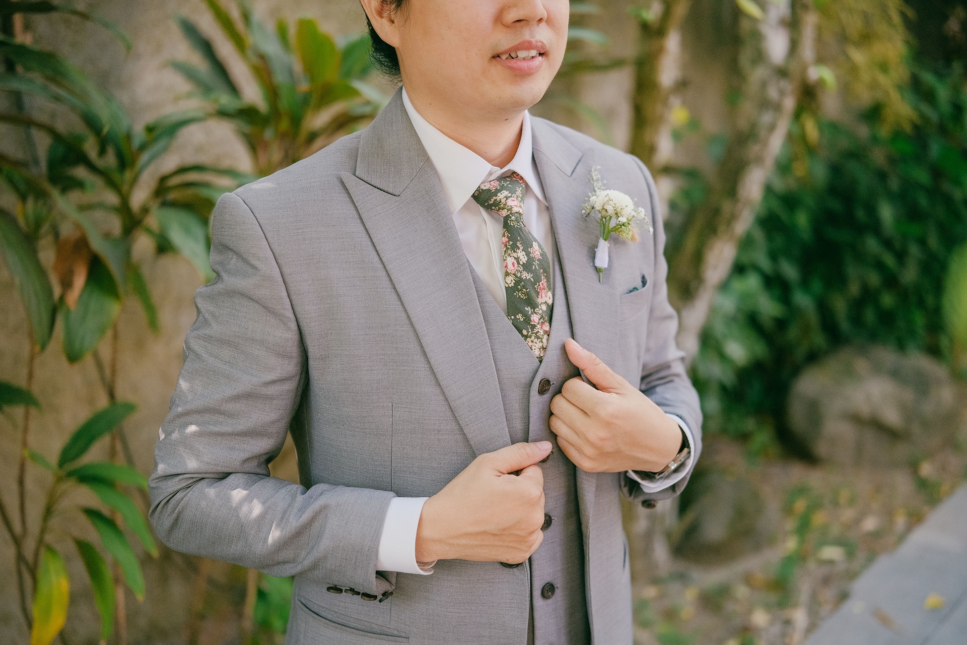 Close Up of Asian Groom in Elegant Gray Suit Standing in Lush Green Garden Holding his Jacket